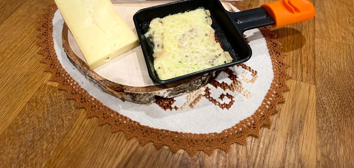 Raclette-Pfännchen mit geschmolzenem Landgraf neben Käsescheiben auf einem Holzbrett